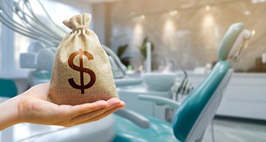 A hand holding a money bag in front of a teal dental chair